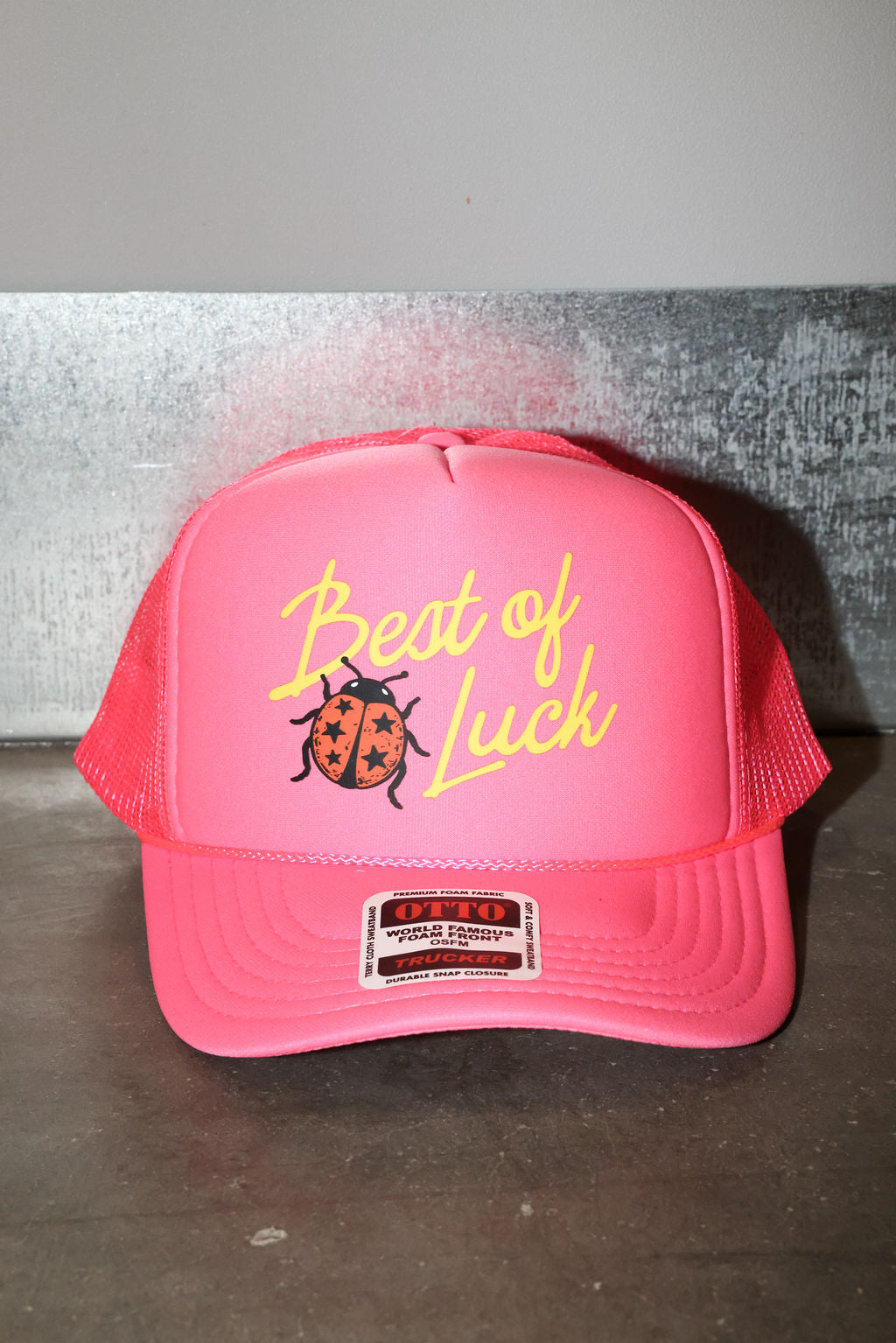 A bright pink trucker hat with mesh back panels, featuring a yellow embroidered 'Best of Luck' script and a ladybug graphic on the front. A sticker is visible on the brim, and the hat is photographed against a gray textured background