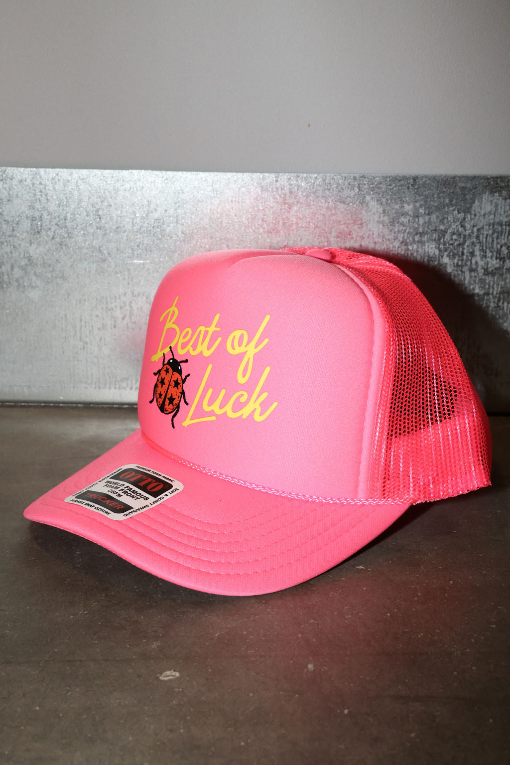 A bright pink trucker hat with a curved brim, featuring yellow embroidered text reading 'Best of Luck' and a small ladybug graphic on the front panel. The back of the cap is made of pink mesh