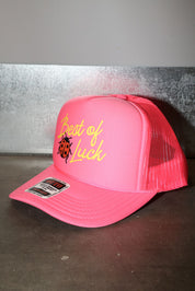 A bright pink trucker hat with a curved brim, featuring yellow embroidered text reading 'Best of Luck' and a small ladybug graphic on the front panel. The back of the cap is made of pink mesh