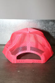 A bright neon pink trucker hat viewed from the back, featuring a solid pink crown and mesh back panels, resting on a dark surface against a gray wall