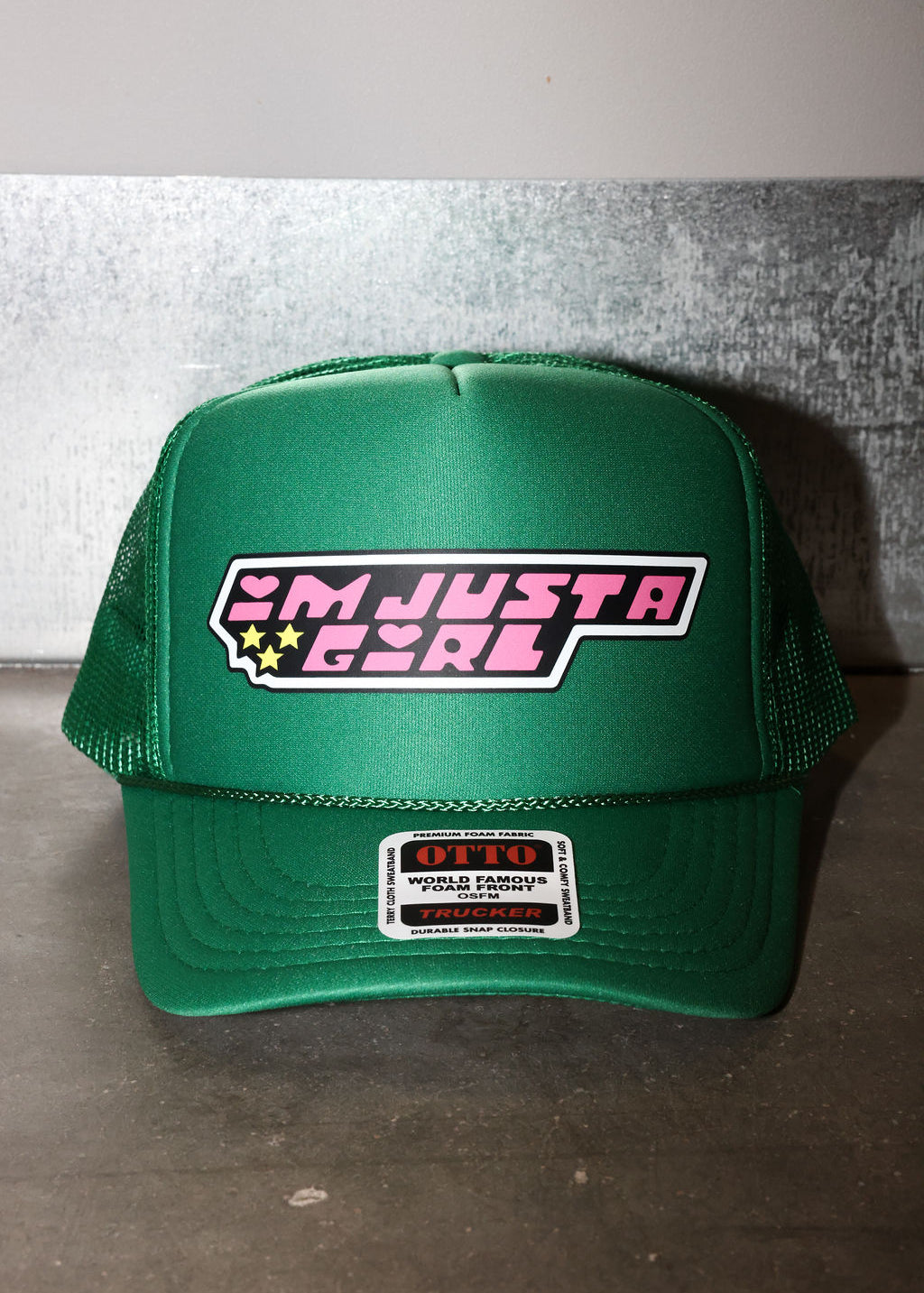 A green trucker hat with a mesh back and a front patch that reads 'I'M JUST A GIRL' in pink and white with small yellow stars. A smaller 'OTTO' brand patch is visible on the brim