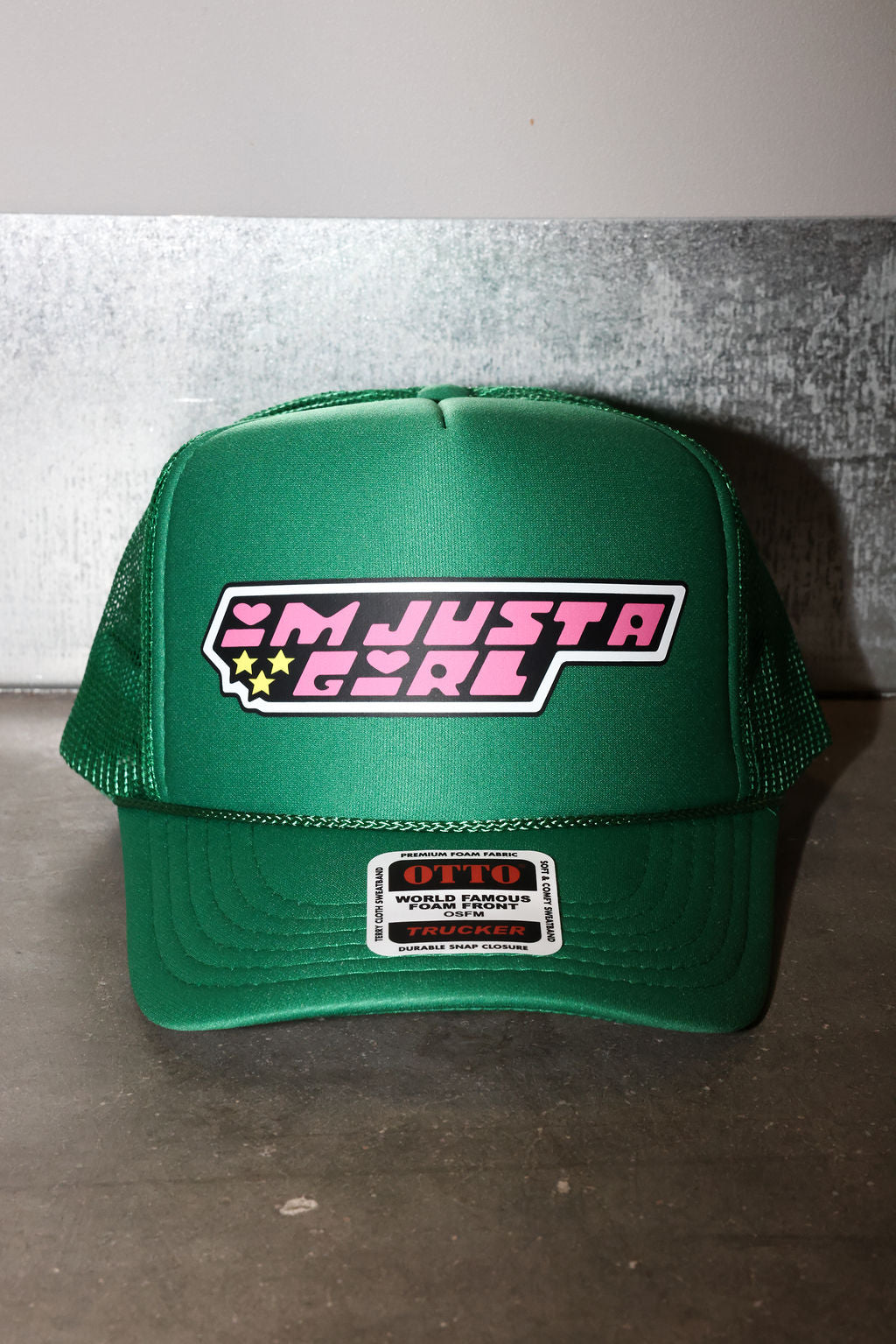 A green trucker hat with a mesh back and a front patch that reads 'I'M JUST A GIRL' in pink and white with small yellow stars. A smaller 'OTTO' brand patch is visible on the brim