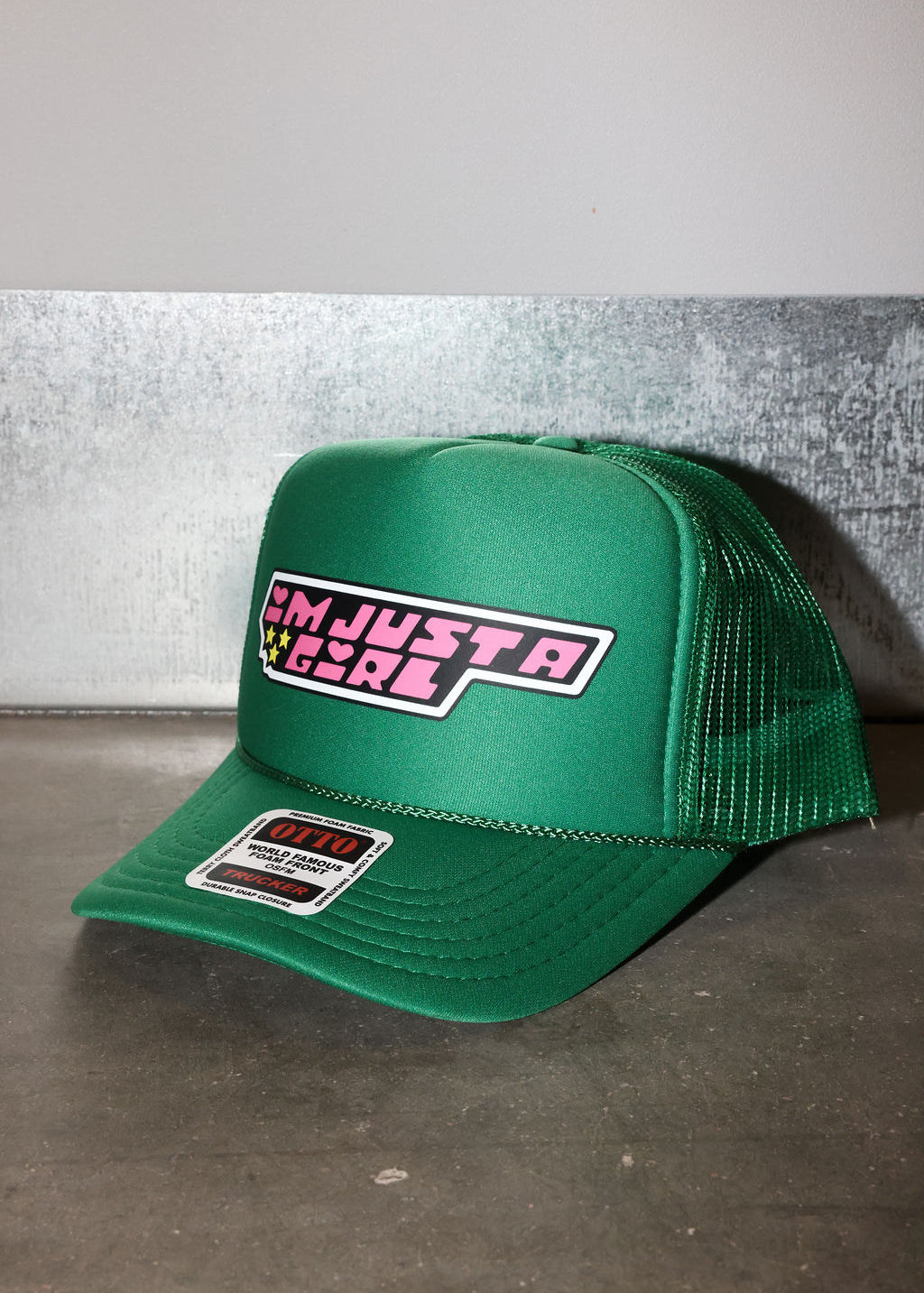 A green trucker hat with a curved brim, featuring a front patch that reads 'I'M JUST A GIRL' in pink and white, and a brand sticker on the brim