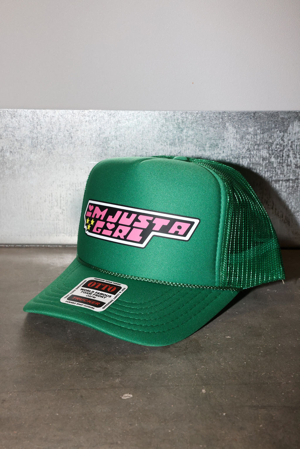 A green trucker hat with a curved brim, featuring a front patch that reads 'I'M JUST A GIRL' in pink and white, and a brand sticker on the brim