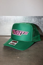 A green trucker hat with a curved brim, featuring a front patch that reads 'I'M JUST A GIRL' in pink and white, and a brand sticker on the brim