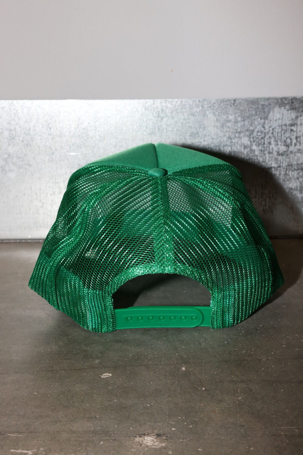 A bright green trucker-style cap with a mesh back, photographed from the rear against a gray textured wall and concrete floor