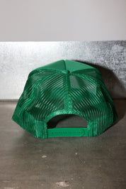 A bright green trucker-style cap with a mesh back, photographed from the rear against a gray textured wall and concrete floor