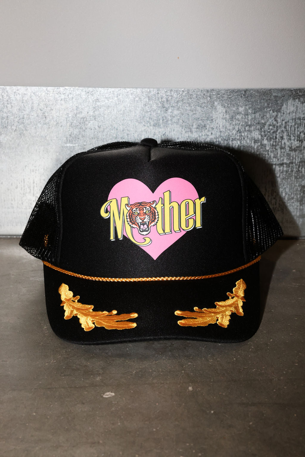 A black trucker-style cap with a pink heart graphic on the front, the word 'Mother' in yellow script with a tiger face in the center, and gold embroidered wing-like details on the brim