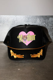 A black trucker-style cap with a pink heart graphic on the front, the word 'Mother' in yellow script with a tiger face in the center, and gold embroidered wing-like details on the brim