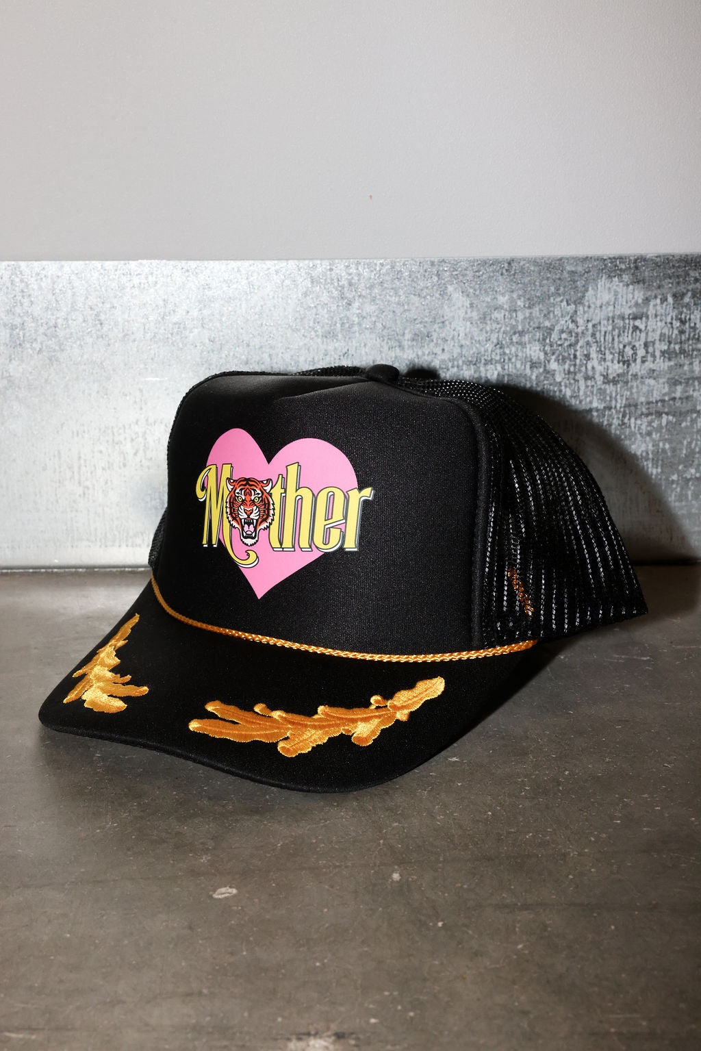 A black trucker-style cap with a pink heart graphic on the front, the word 'Mother' in yellow script with a tiger face in the center, and gold embroidered wing-like details on the brim, side view