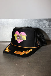 A black trucker-style cap with a pink heart graphic on the front, the word 'Mother' in yellow script with a tiger face in the center, and gold embroidered wing-like details on the brim, side view