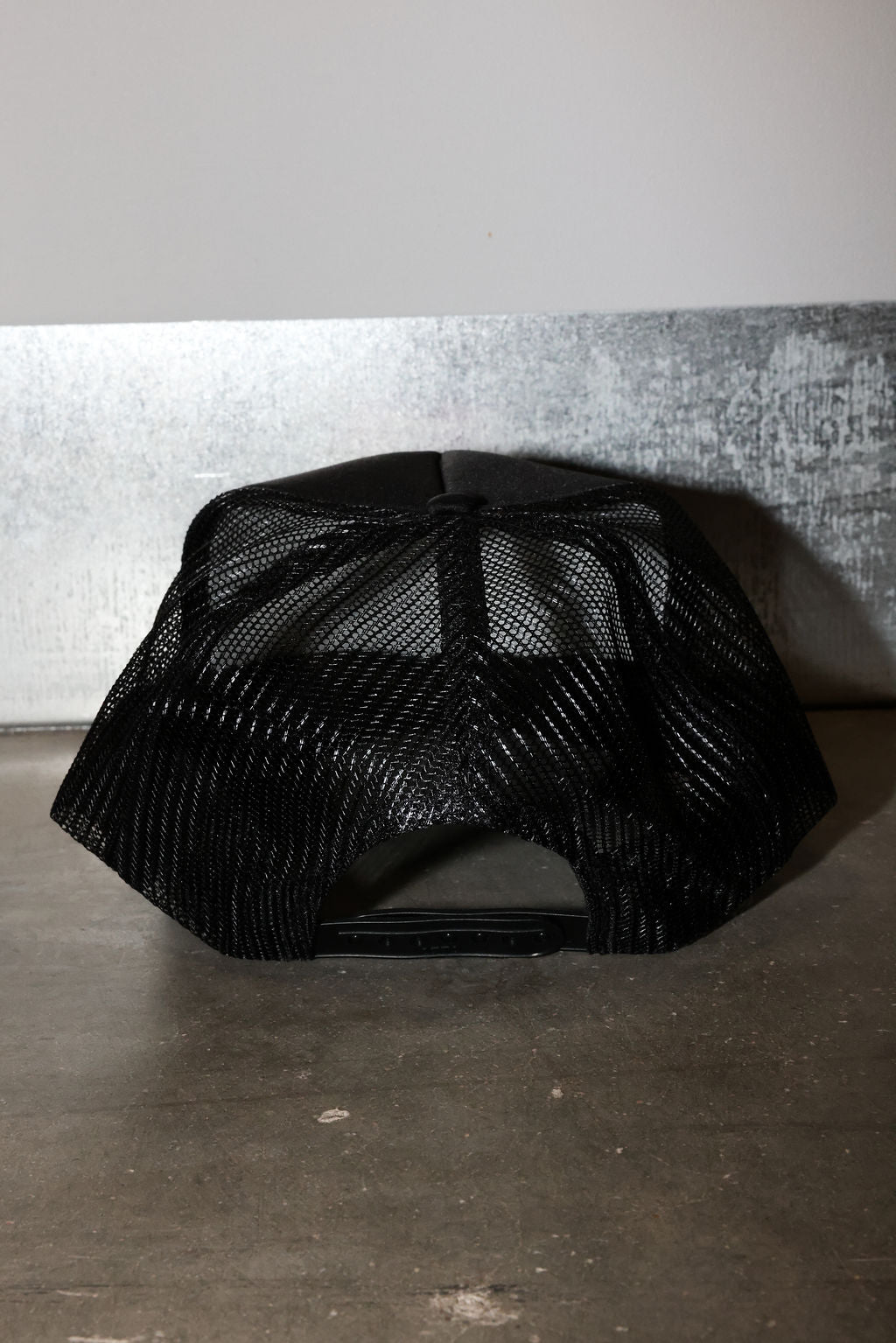 A black trucker-style cap with a black mesh back, photographed on a dark concrete floor against a gray wall