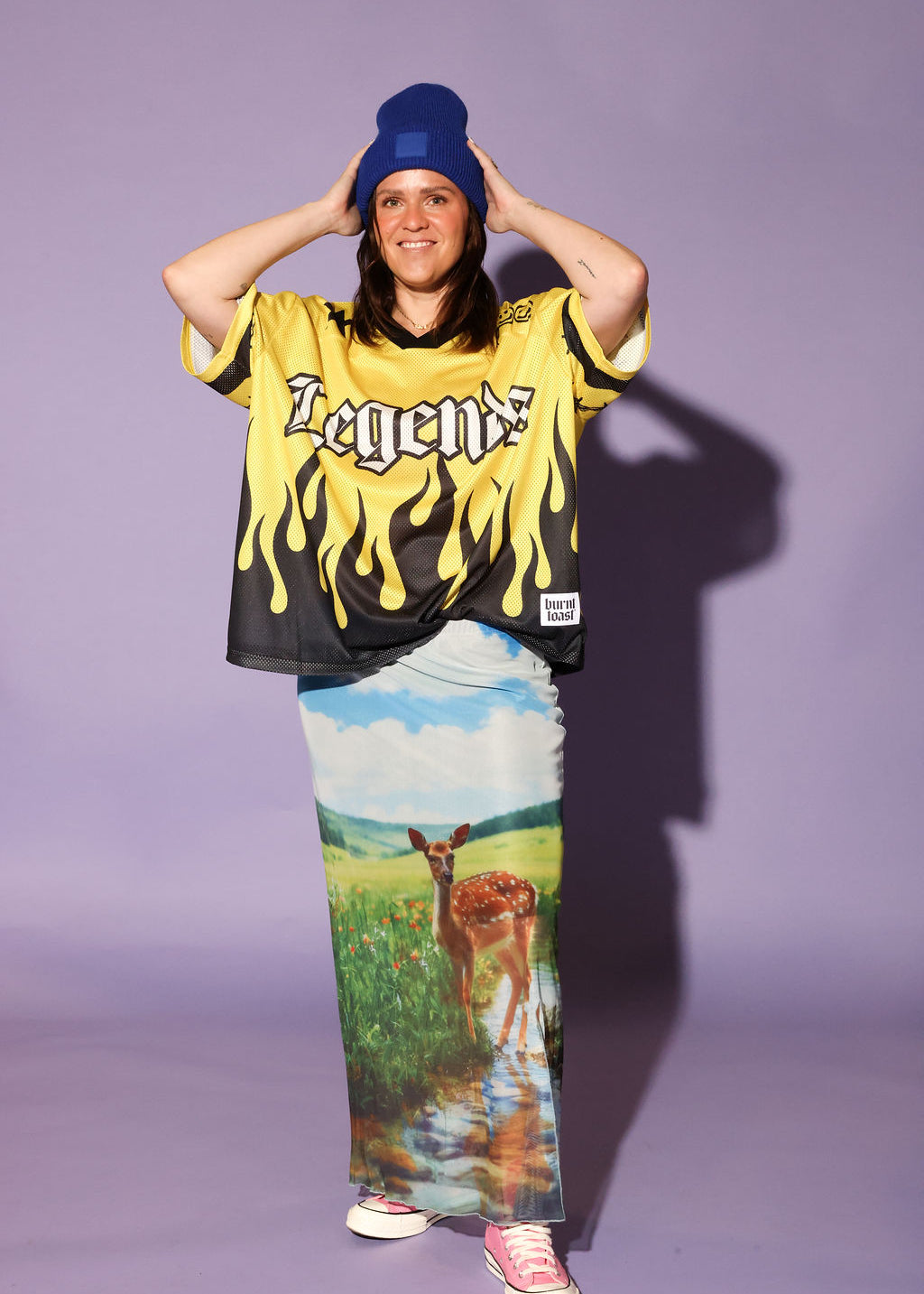 A model stands against a solid lavender studio background wearing a yellow and black flame-print graphic t-shirt with 'Legend' written on the chest, a long printed maxi skirt featuring a deer in a field, a blue beanie, and pink sneakers, frontal view
