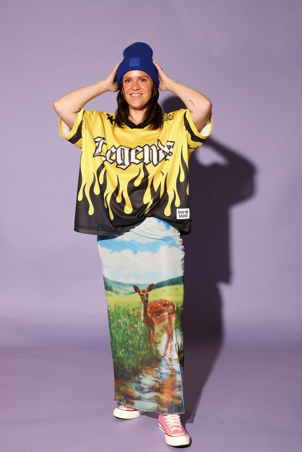 A model stands against a solid lavender studio background wearing a yellow and black flame-print graphic t-shirt with 'Legend' written on the chest, a long printed maxi skirt featuring a deer in a field, a blue beanie, and pink sneakers, frontal view