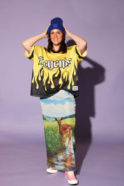A model stands against a solid lavender studio background wearing a yellow and black flame-print graphic t-shirt with 'Legend' written on the chest, a long printed maxi skirt featuring a deer in a field, a blue beanie, and pink sneakers, frontal view