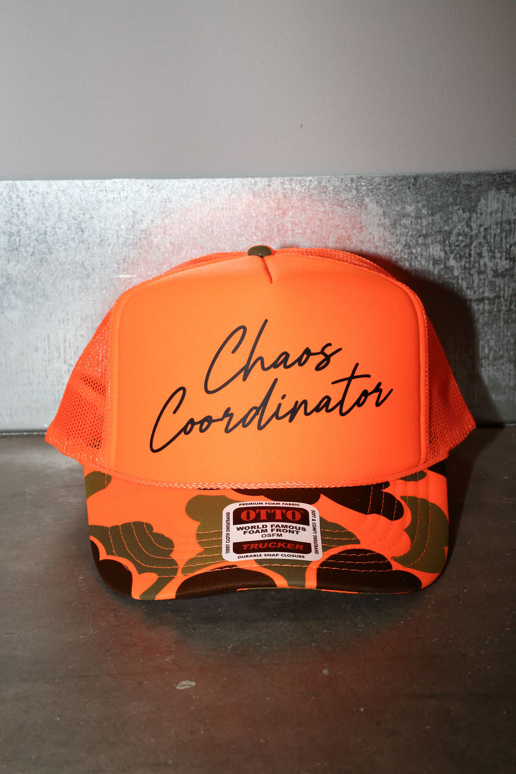 An orange trucker hat with 'Chaos Coordinator' written in black script on the front. The brim features a green and orange camouflage pattern with a white and patch