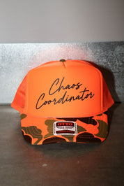 An orange trucker hat with 'Chaos Coordinator' written in black script on the front. The brim features a green and orange camouflage pattern with a white and patch