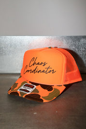 An orange trucker hat with a camouflage-patterned visor and orange mesh back, featuring the words 'Chaos Coordinator' in black sticker on the front panel