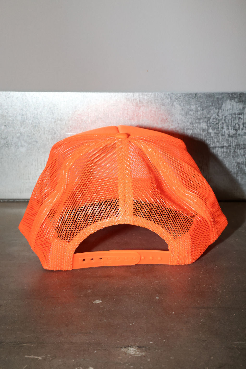 A bright neon orange trucker hat with a mesh back, photographed from the back on a dark grey surface against a gray wall