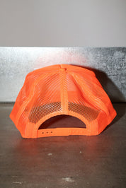 A bright neon orange trucker hat with a mesh back, photographed from the back on a dark grey surface against a gray wall