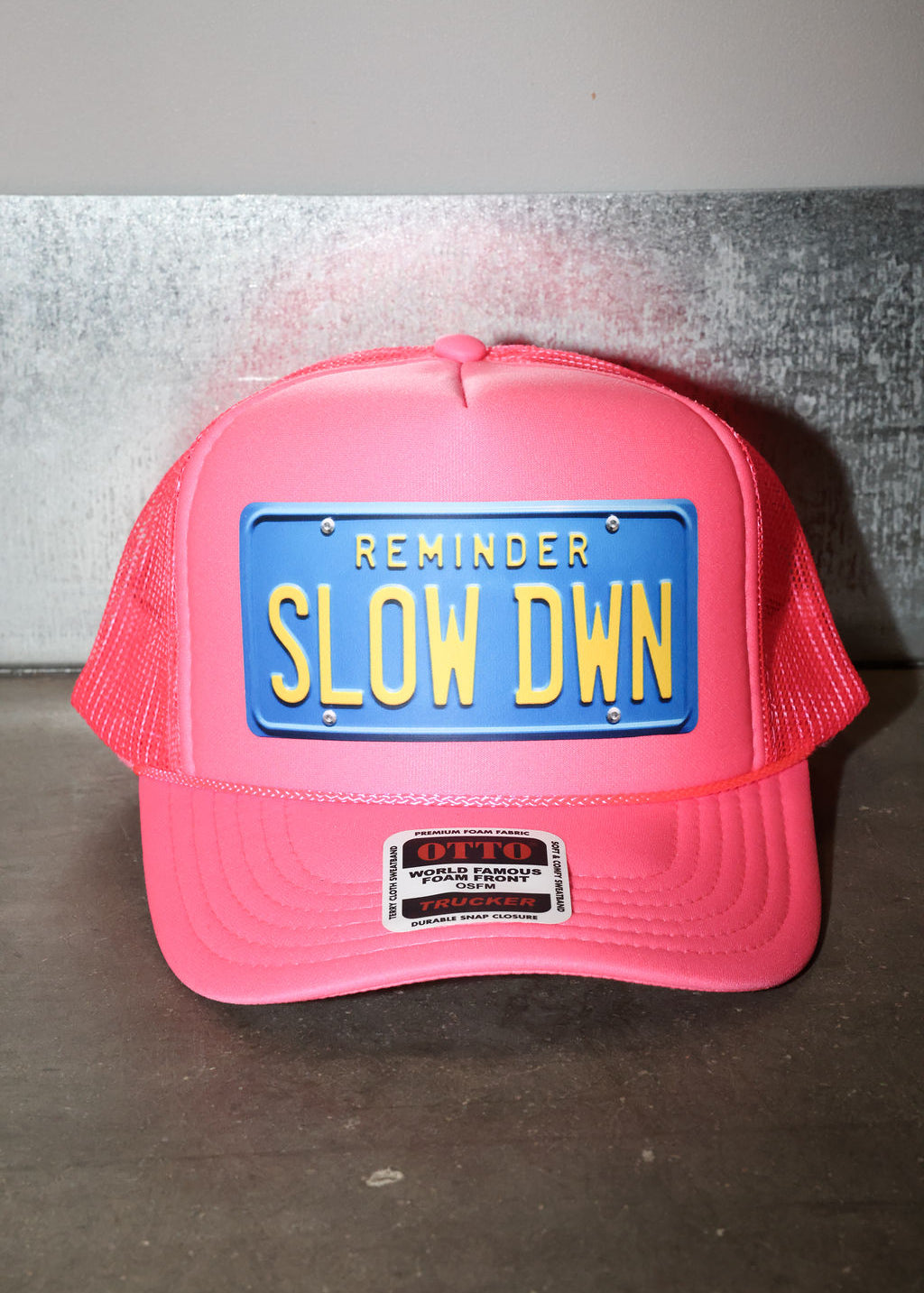 Pink trucker hat with blue license plate and text that read 'REMINDER SLOW DOWN' in yellow, flat lay frontal view