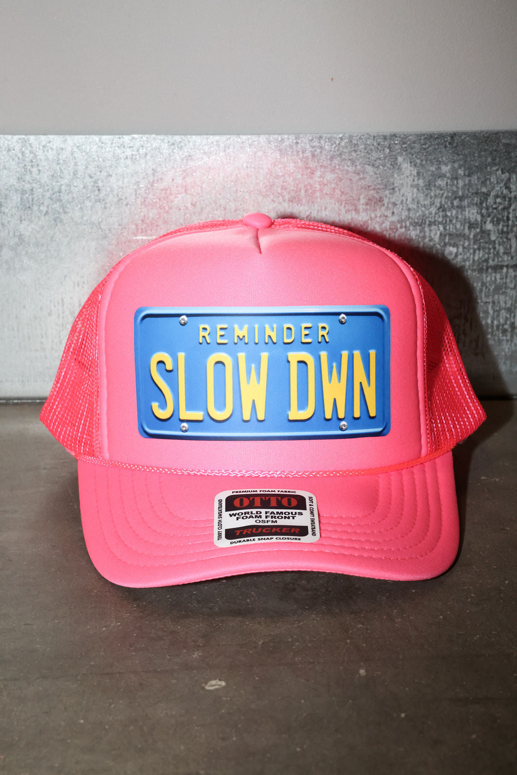 Pink trucker hat with blue license plate and text that read 'REMINDER SLOW DOWN' in yellow, flat lay frontal view
