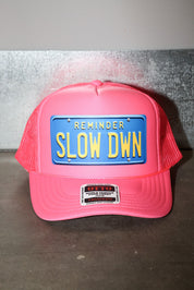 Pink trucker hat with blue license plate and text that read 'REMINDER SLOW DOWN' in yellow, flat lay frontal view