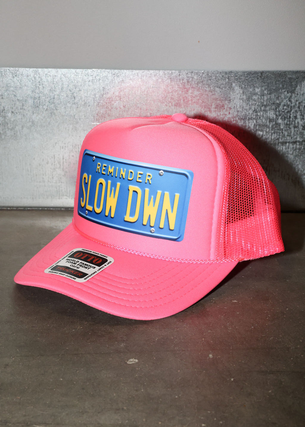 Pink trucker hat with a blue license plate reading 'SLOW DOWN' on a gray background, side view