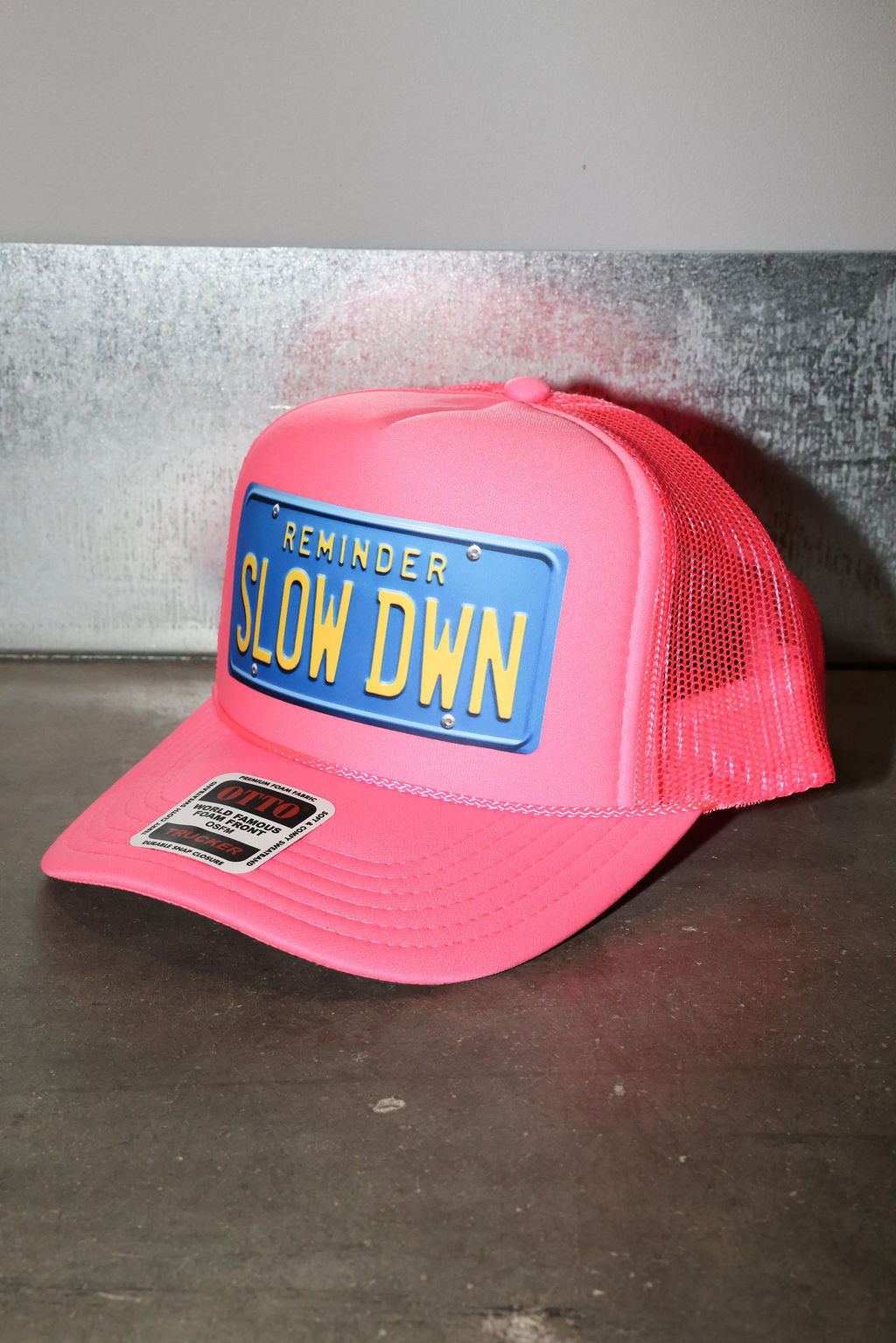 Pink trucker hat with a blue license plate reading 'SLOW DOWN' on a gray background, side view
