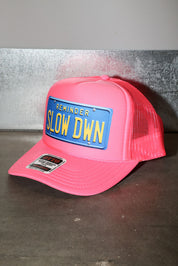 Pink trucker hat with a blue license plate reading 'SLOW DOWN' on a gray background, side view