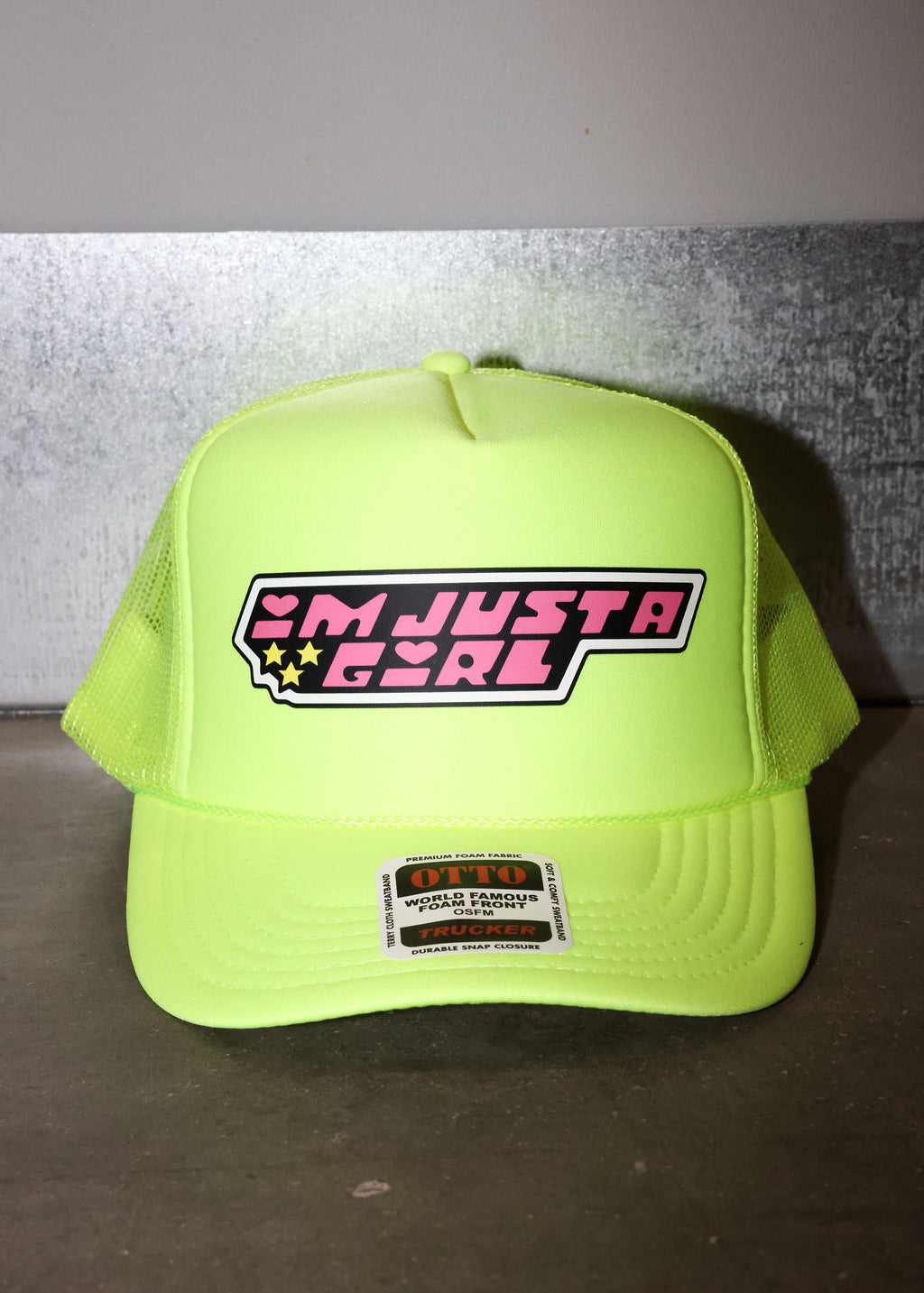 A neon yellow-green trucker hat with a 'I'M JUST A GIRL' graphic on the front panel, displayed on a dark surface against a gray wall