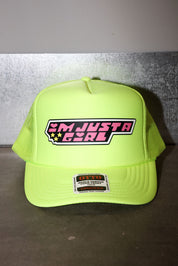 A neon yellow-green trucker hat with a 'I'M JUST A GIRL' graphic on the front panel, displayed on a dark surface against a gray wall