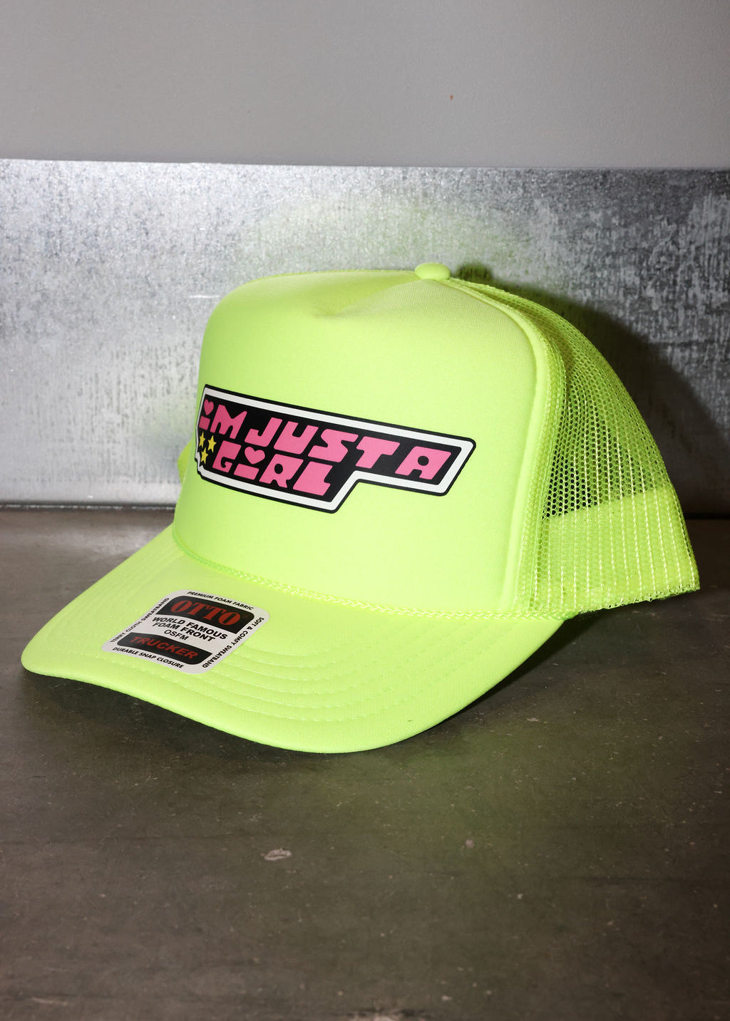 A neon yellow-green trucker hat with mesh back panels and a front patch reading 'I'M JUST A GIRL'. The hat is photographed on a dark surface against a gray wall