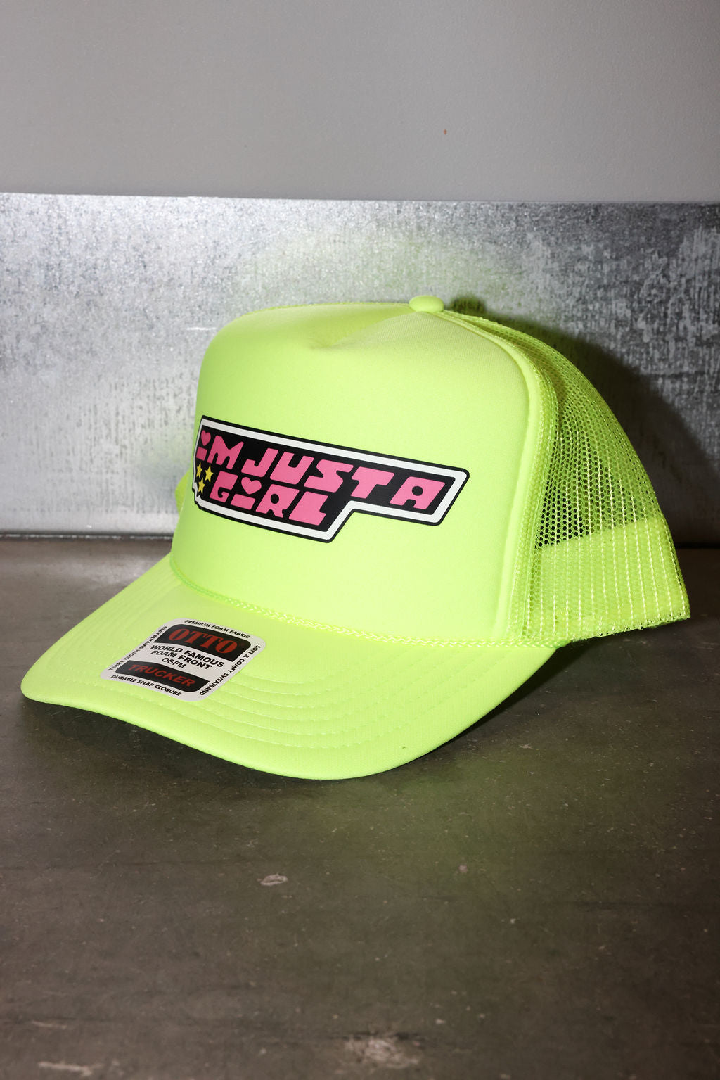 A neon yellow-green trucker hat with mesh back panels and a front patch reading 'I'M JUST A GIRL'. The hat is photographed on a dark surface against a gray wall