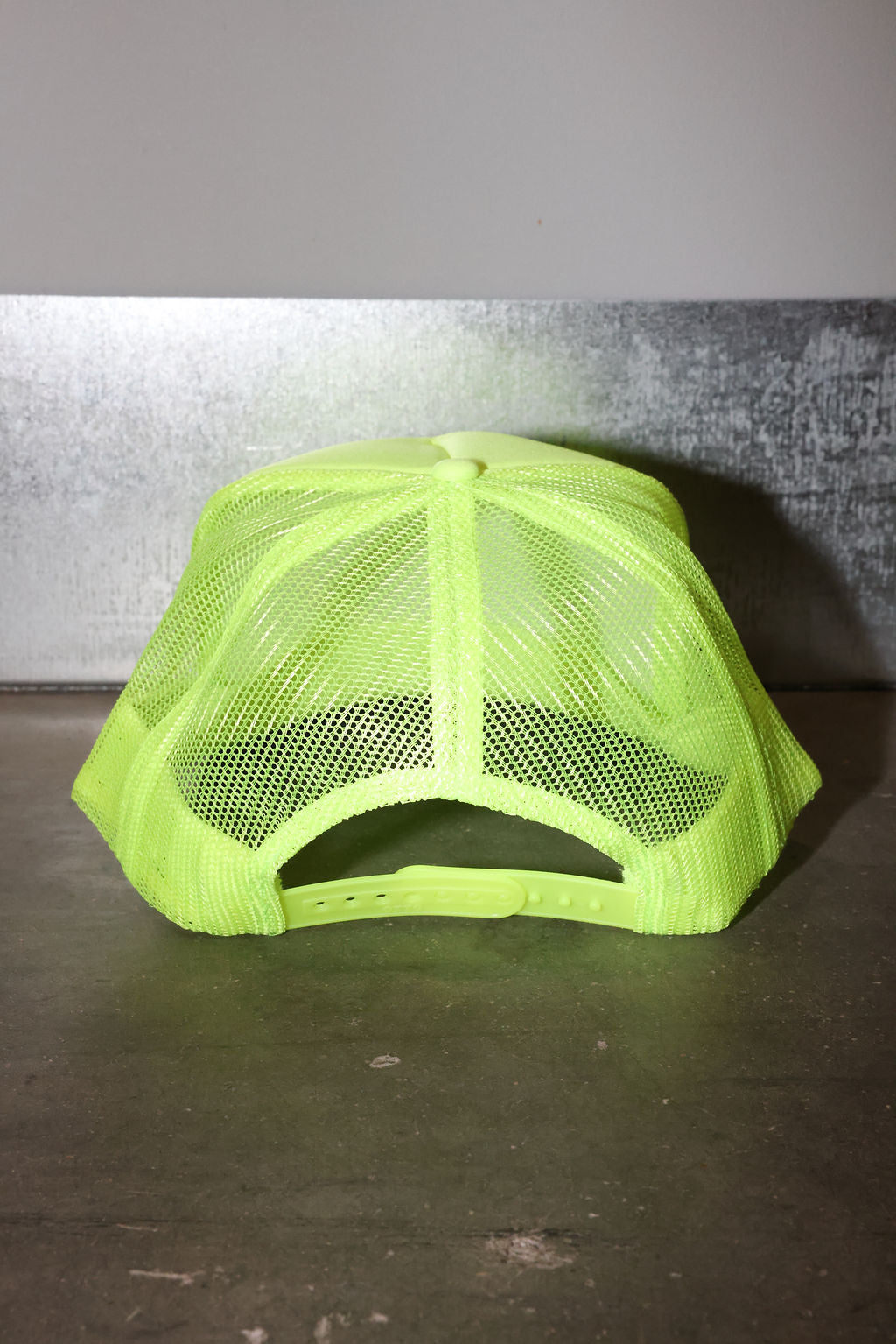 A neon yellow-green trucker hat with a mesh back, photographed from the back against a textured gray wall and dark surface