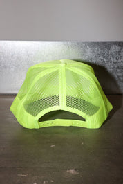 A neon yellow-green trucker hat with a mesh back, photographed from the back against a textured gray wall and dark surface