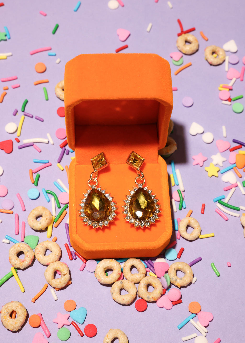 Open orange jewelry box with amber-colored earrings on a colorful sprinkles and cereal background