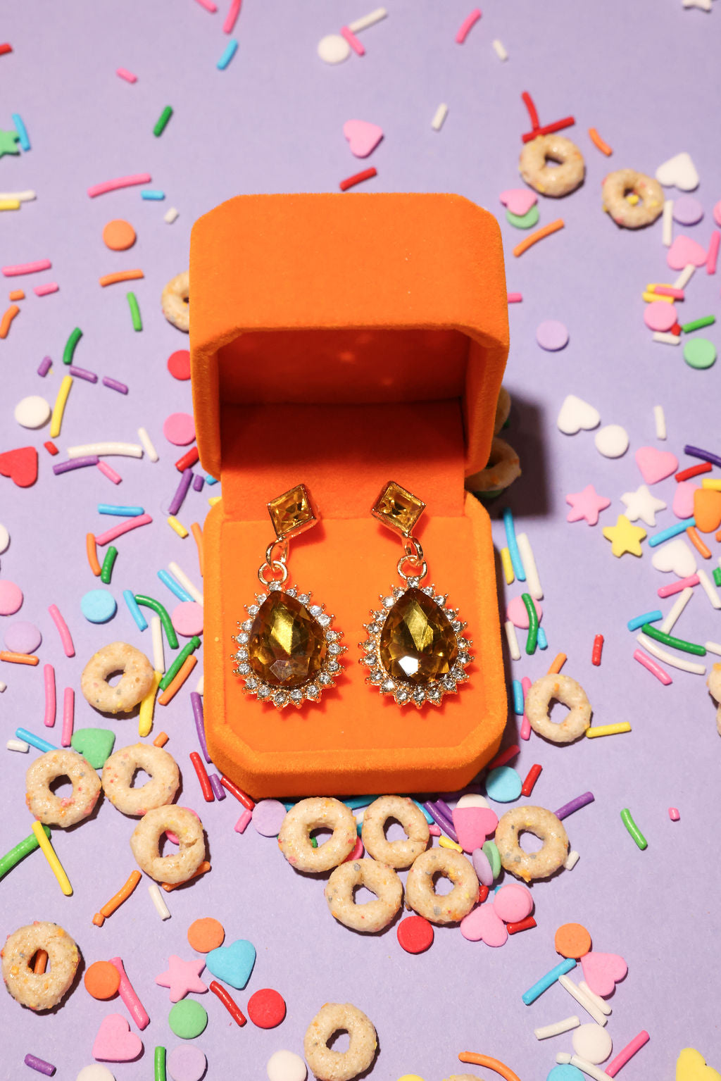 Open orange jewelry box with amber-colored earrings on a colorful sprinkles and cereal background