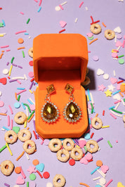 Open orange jewelry box with amber-colored earrings on a colorful sprinkles and cereal background