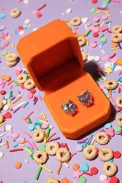 Open orange jewelry box with heart-shaped earrings on a colorful cereal-themed background