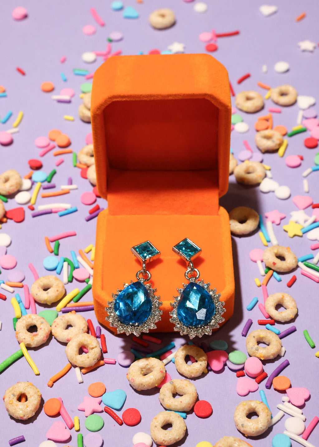 Blue earrings in an open orange jewelry box surrounded by colorful sprinkles and donuts on a purple background