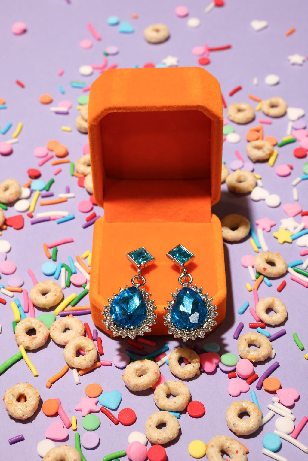 Blue earrings in an open orange jewelry box surrounded by colorful sprinkles and donuts on a purple background