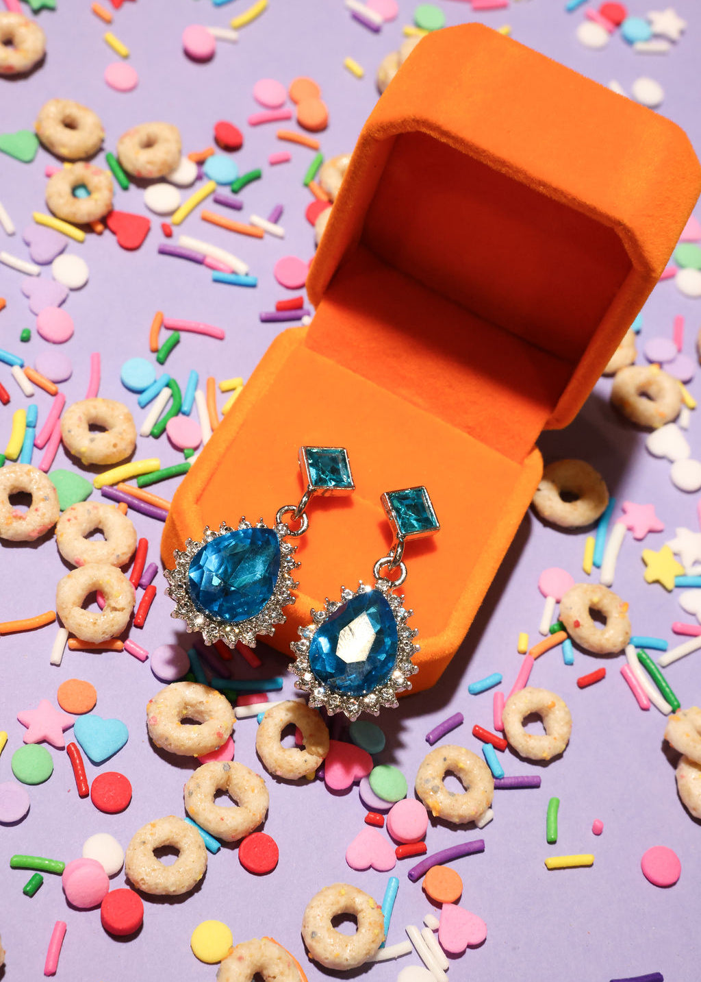 Blue gemstone earrings in an orange jewelry box with colorful cereal and sprinkles on a purple background