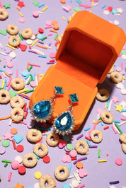 Blue gemstone earrings in an orange jewelry box with colorful cereal and sprinkles on a purple background
