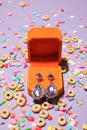 Earrings in an orange box surrounded by colorful sprinkles and donut-shaped candies on a purple background