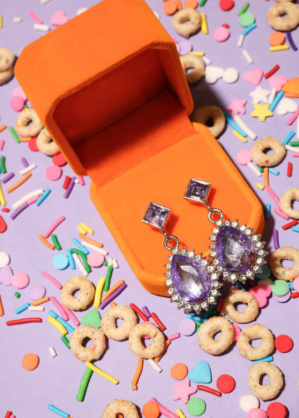 Purple teardrop earrings in an orange jewelry box with colorful sprinkles and cereal on a purple background