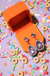 Purple teardrop earrings in an orange jewelry box with colorful sprinkles and cereal on a purple background