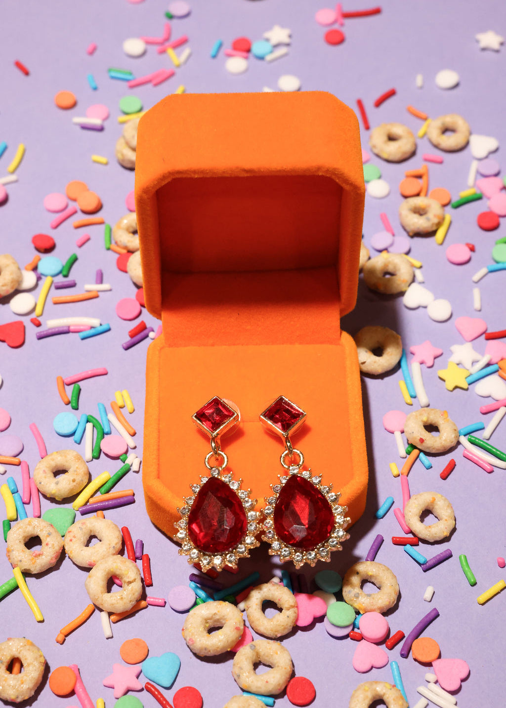 Red earrings in an open orange jewelry box with colorful sprinkles and cereal on a purple background