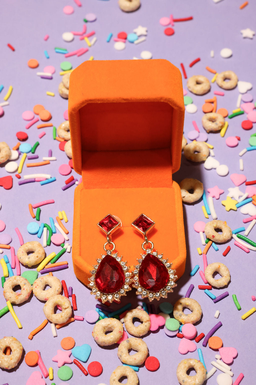 Red earrings in an open orange jewelry box with colorful sprinkles and cereal on a purple background