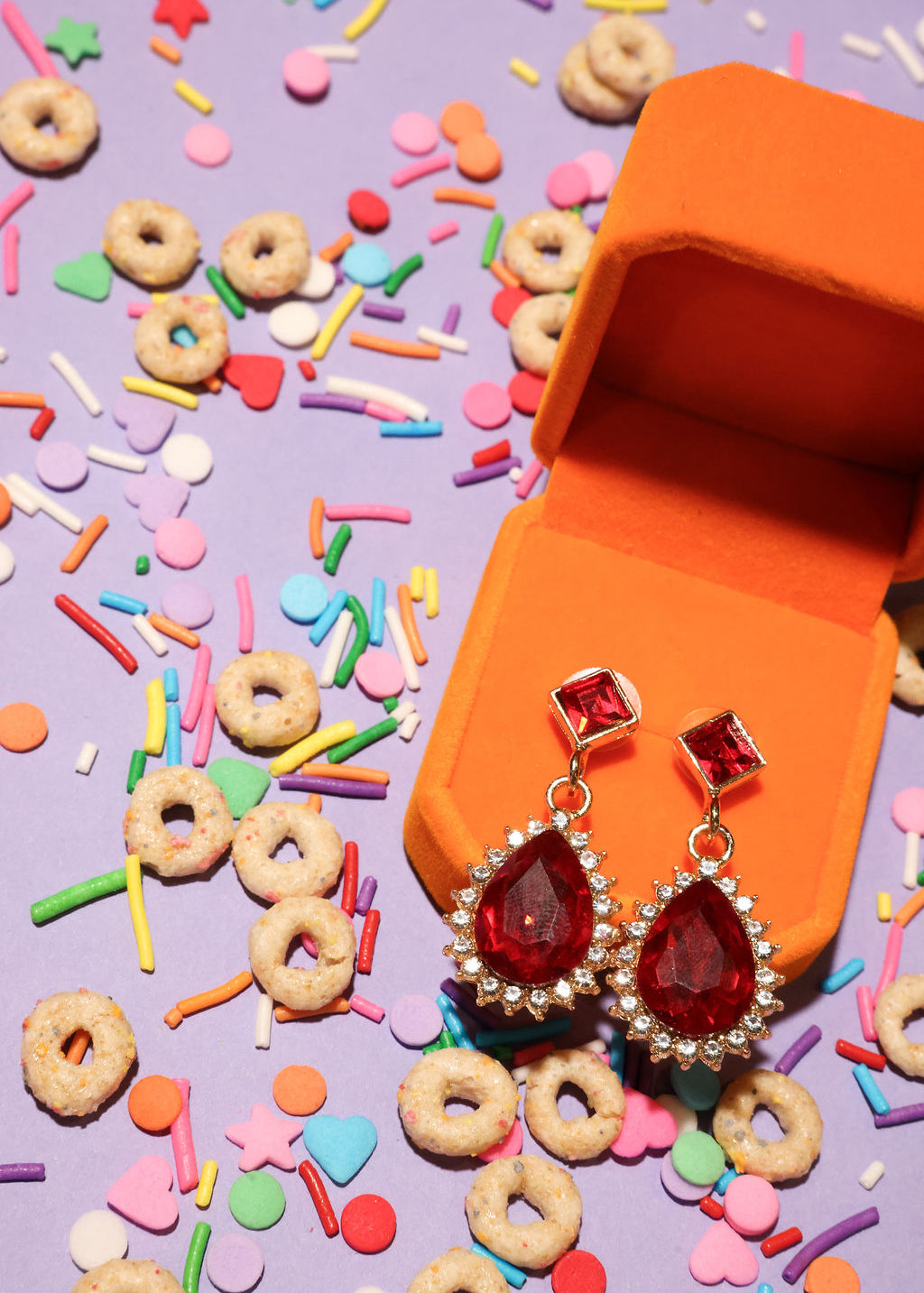 Red teardrop earrings in an orange jewelry box with colorful sprinkles and cereal on a purple background